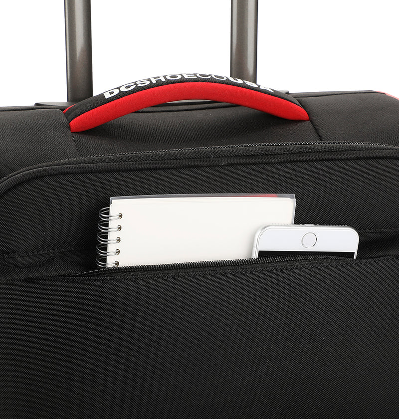 A close-up of the DC Shoes DC Runner Carry-On shows its black design with a red and black handle. A spiral notebook and smartphone peek out from the front zippered pocket.