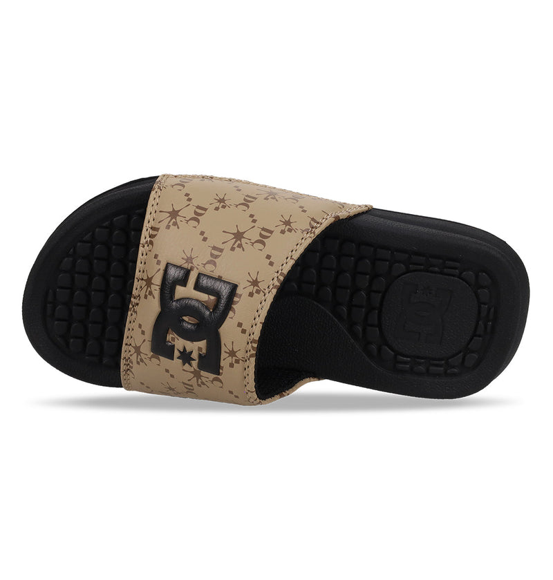 The DC Shoes Kids' Bolsa Slides feature a beige and black design with abstract star patterns on the strap, a bold black logo, and a textured black footbed—perfect for any kids' slide sandal collection.