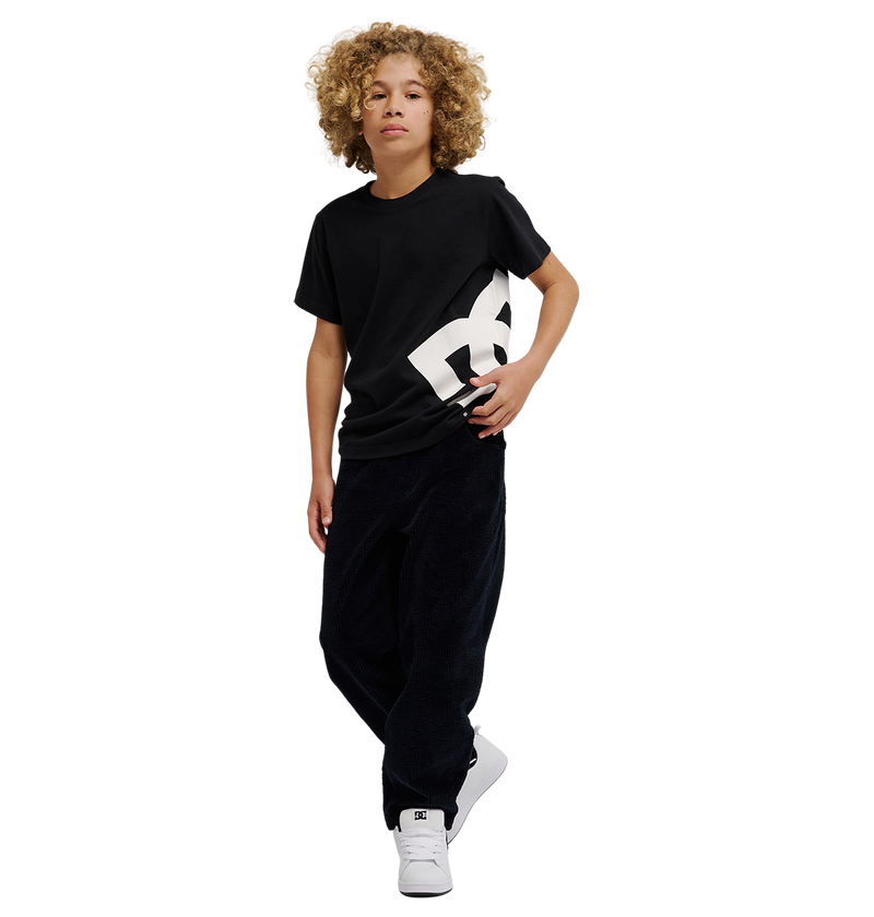 A young person with curly blond hair wears the DC Shoes DC Lanai Tee Youth, a black relaxed-fit graphic skate shirt with a bold white logo, along with loose black pants and white sneakers, standing against a plain white background.