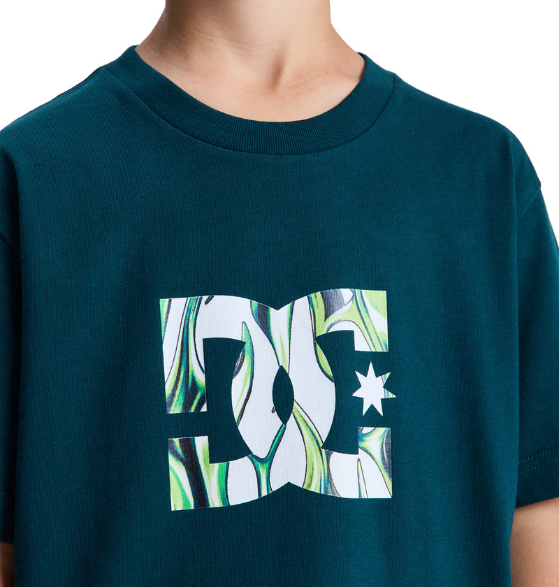 Someone wearing the Reebok DC Star Filled Ss Boy T-shirt in teal, featuring a large abstract DC Shoes logo with green, white, and black swirls on the chest. The photo is cropped to show only the shirt and neck.