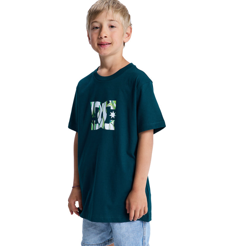 A young boy with blonde hair wears the Reebok DC Star Filled Ss Boy t-shirt in dark green with a patterned logo and light denim shorts, smiling at the camera against a white background.