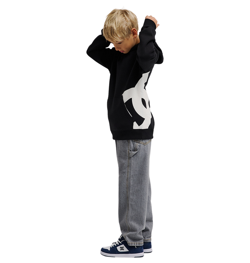 A young boy with blonde hair wears the DC Shoes Lanai Hoodie Youth—black with bold DC graphics—paired with gray jeans and blue sneakers, standing sideways and adjusting his hood for warmth against a white background.