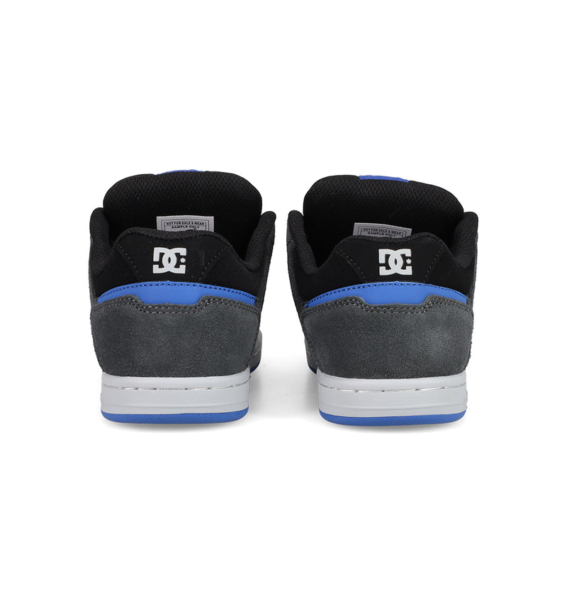A pair of Kids' Stag Shoes by DC Shoes seen from the back, featuring black and grey suede uppers, blue trim, white soles, cupsole construction, and the DC logo on the heel.
