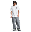A young person with braided hair, wearing a white DC Shoes t-shirt, Men's Baggy Denim Carpenter Pant by DC Shoes, and black and green sneakers stands looking down with one hand in their pocket against a plain white background.