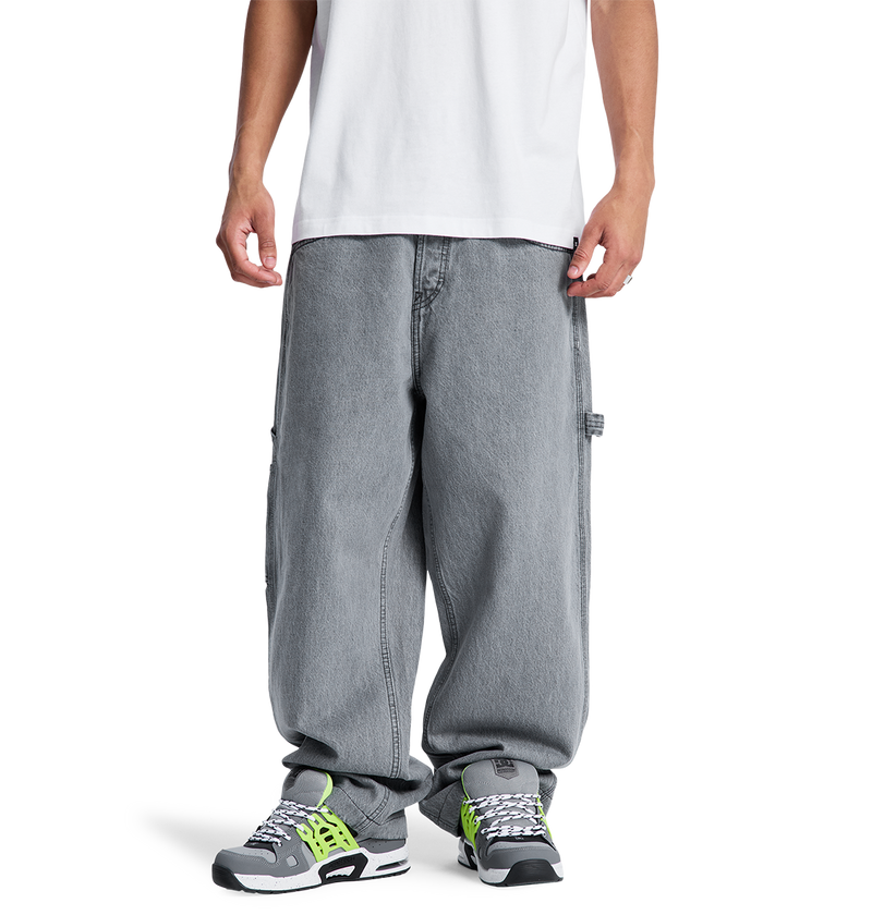 A person stands facing forward, hands relaxed, wearing a white T-shirt and DC Shoes Men's Baggy Denim Carpenter Pant in gray, paired with gray sneakers featuring green and black accents. Photo is cropped just below the shoulders.