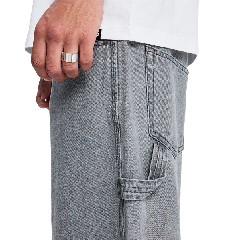 A close-up of a hand wearing a silver ring rests near the pocket of DC Shoes Men's Baggy Denim Carpenter Pant in light gray with visible stitching, shown next to a white shirt.