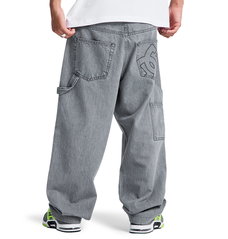 A person stands facing away, wearing DC Shoes Men's Baggy Denim Carpenter Pant with large pockets and a white graphic on the back pocket, styled with a white shirt and chunky sneakers featuring neon green accents.