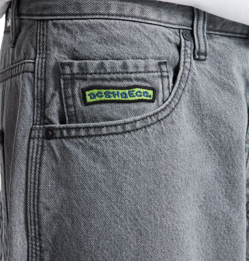Close-up of the front pocket of the Men's Baggy Denim Carpenter Pant by DC Shoes, showing a small patch above the pocket with "DC SHO ECO" in green and blue text. A white shirt can be seen above the waistband.