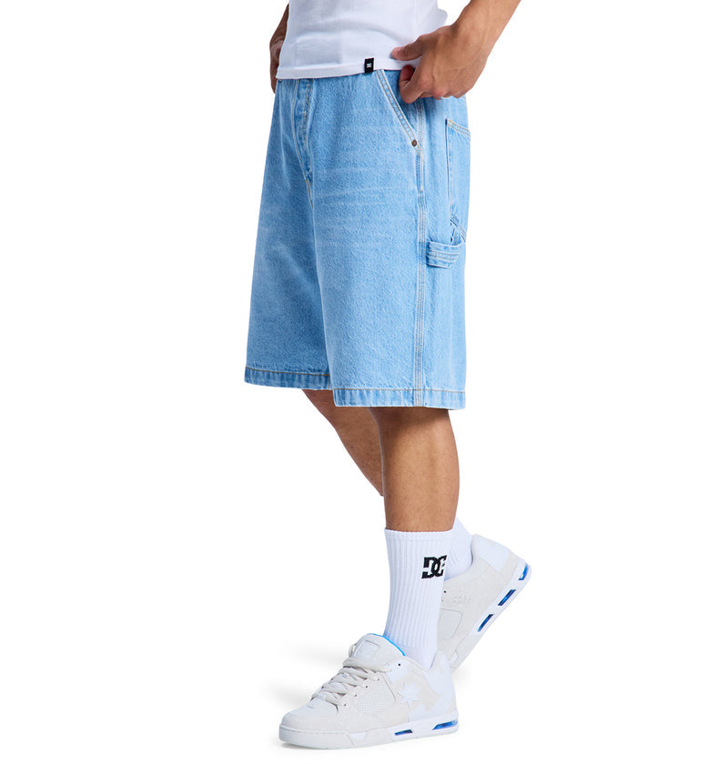Someone wearing Reebok Baggy Denim Carpenter Shorts, a white tee, white DC socks, and white sneakers with blue details. Their hands are in the shorts' pockets, showing only their lower body.