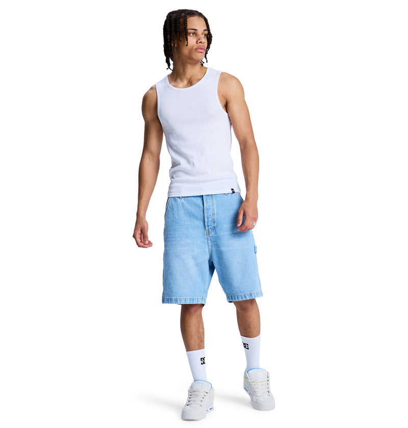 A person wearing a white tank top, Reebok Baggy Denim Carpenter Short in light blue, white socks, and white sneakers stands against a plain white background.
