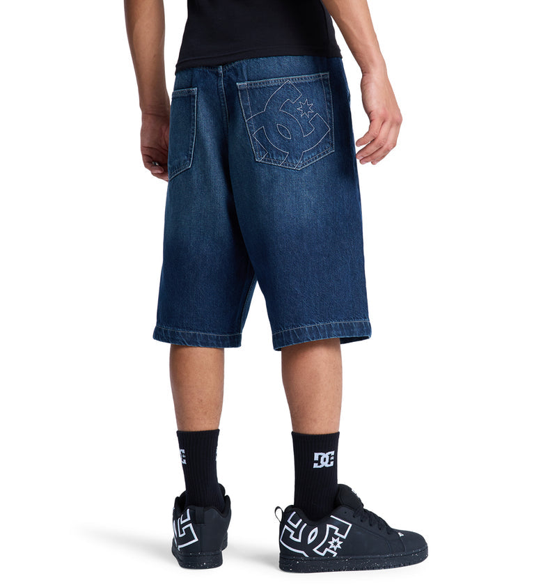 A person in Reebok Baggy Denim Jort, featuring a knee-length fit and blue wash, paired with black socks and black skate shoes, stands against a white background.