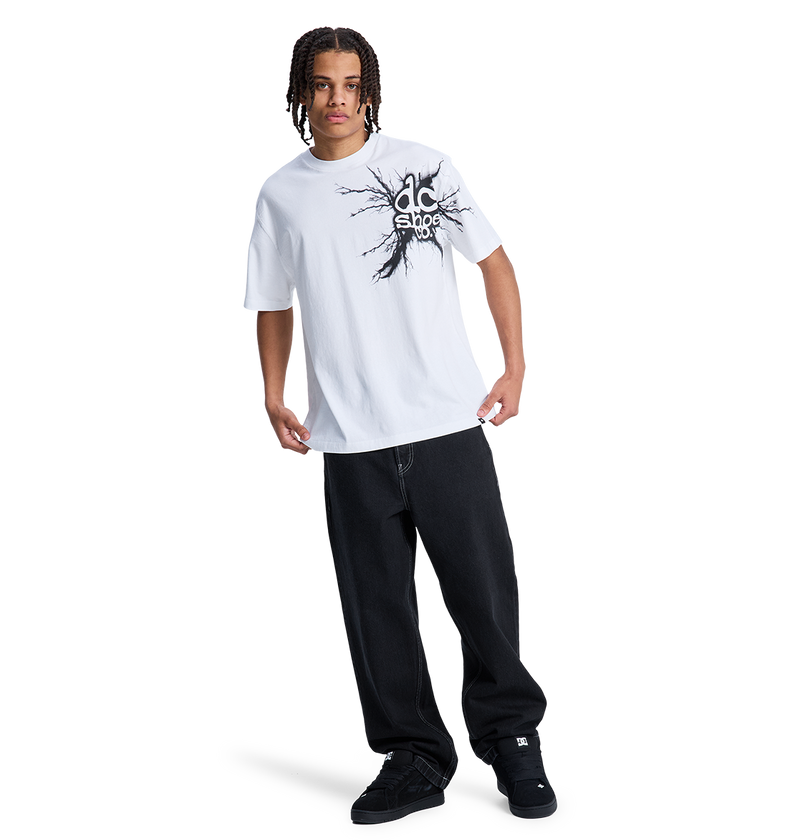 A person with braided hair wears a white graphic tee, black DC Shoes sneakers, and the Men's Baggy Denim Pant by DC Shoes, all set against a plain white background.