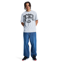 A person with braided hair wears a light gray t-shirt with DISCO and 094, Men's Baggy Denim Pant by DC Shoes in heavyweight denim, and black DC Shoes sneakers, standing against a plain white background.