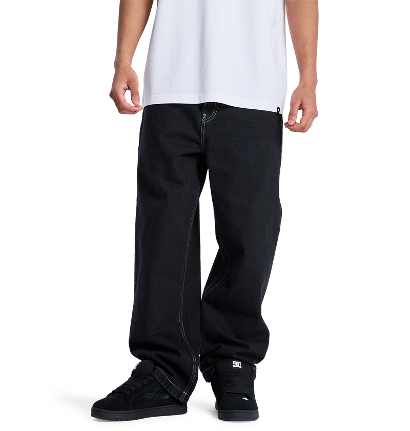 A person stands against a plain white background, wearing DC Shoes Men's Baggy Denim Pant in heavyweight denim with contrast stitching and a loose white T-shirt. The black sneakers are also visible. The image is cropped from the shoulders down.