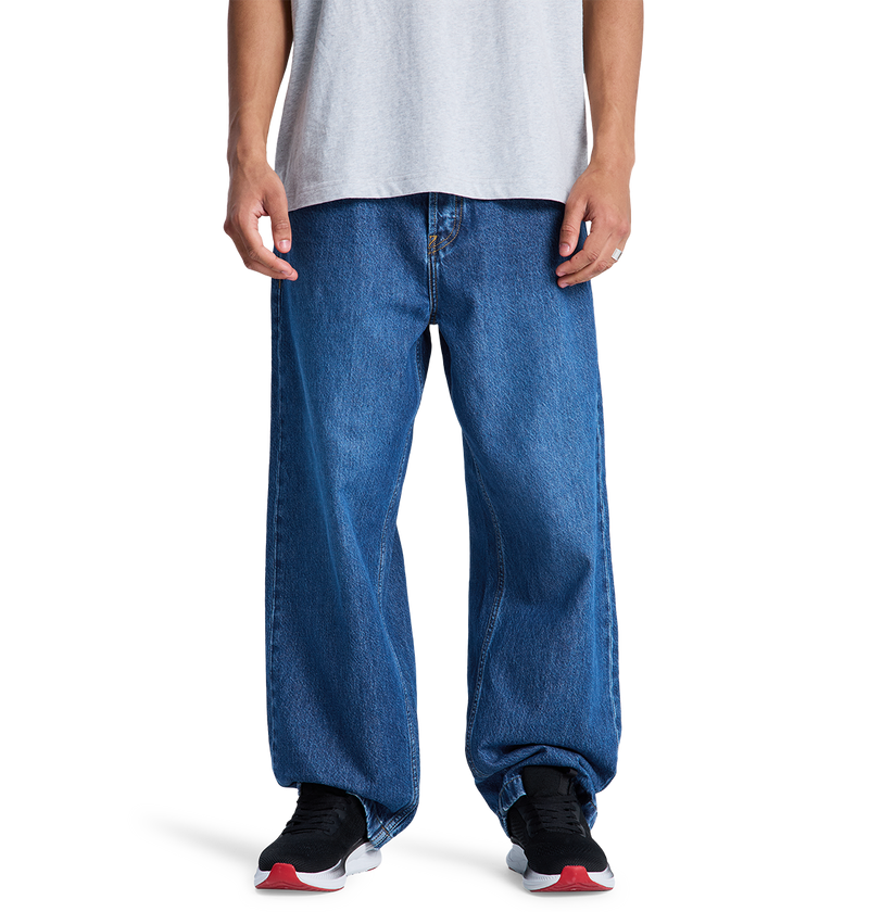 A person wearing DC Shoes' Men's Baggy Denim Pant and black DC sneakers with white soles and red accents stands against a plain white background, paired with a light gray t-shirt.