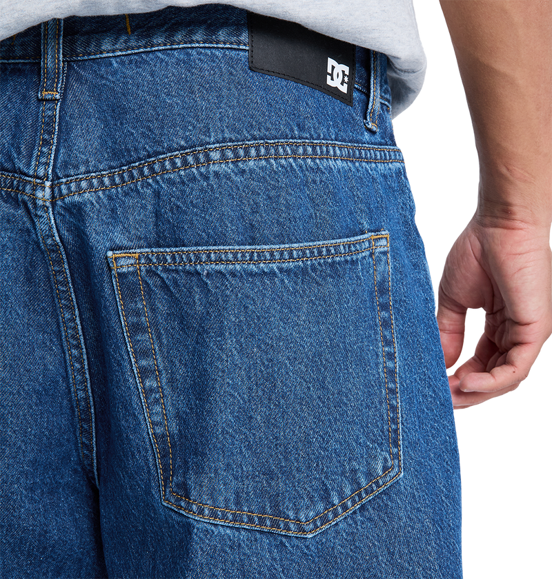 Close-up of the back pocket on DC Shoes Men's Baggy Denim Pant in blue heavyweight denim, showing visible stitching and a black patch with the white DC logo near the waistband. A person's hand rests by their side.