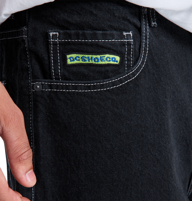 Close-up of a person’s hand near the pocket of DC Shoes Men's Baggy Denim Pant, featuring white stitching and a small DCSHOECO label. They are wearing a white shirt tucked into the heavyweight denim jeans.