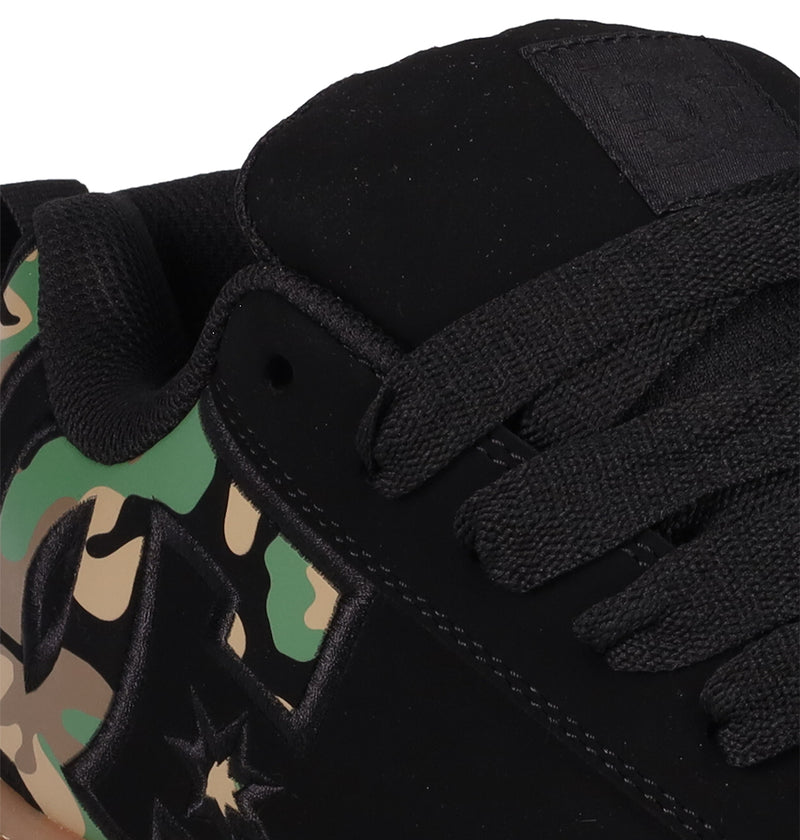 Close-up of DC Shoes Men's Court Graffik: a black sneaker with black laces, green/beige/brown camouflage side panels, and a partially visible star-shaped DC logo on its puffy silhouette.