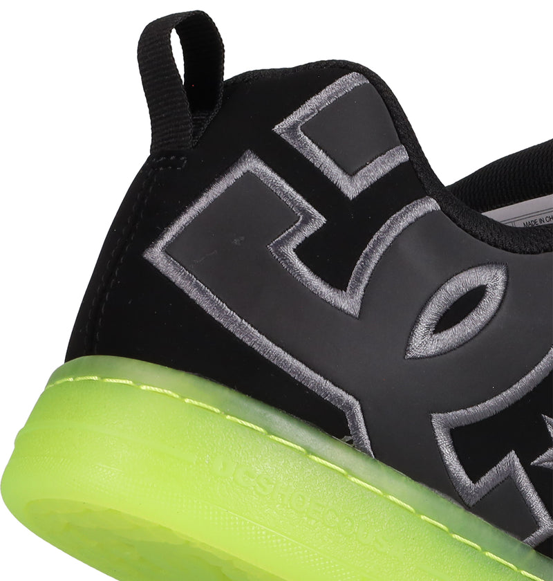 A close-up of the DC Shoes Men's Court Graffik SE showcases its black premium upper, bold silver-outlined lettering, neon green sole, puffy silhouette, and a black pull tab at the heel.