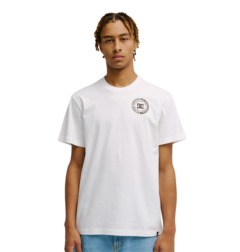 A young man with short, braided hair models the Men's DC Corpo Tee by DC Shoes—a relaxed fit, plain white t-shirt with a small black DC logo and circular emblem on the left chest—paired with light blue jeans against a white background.