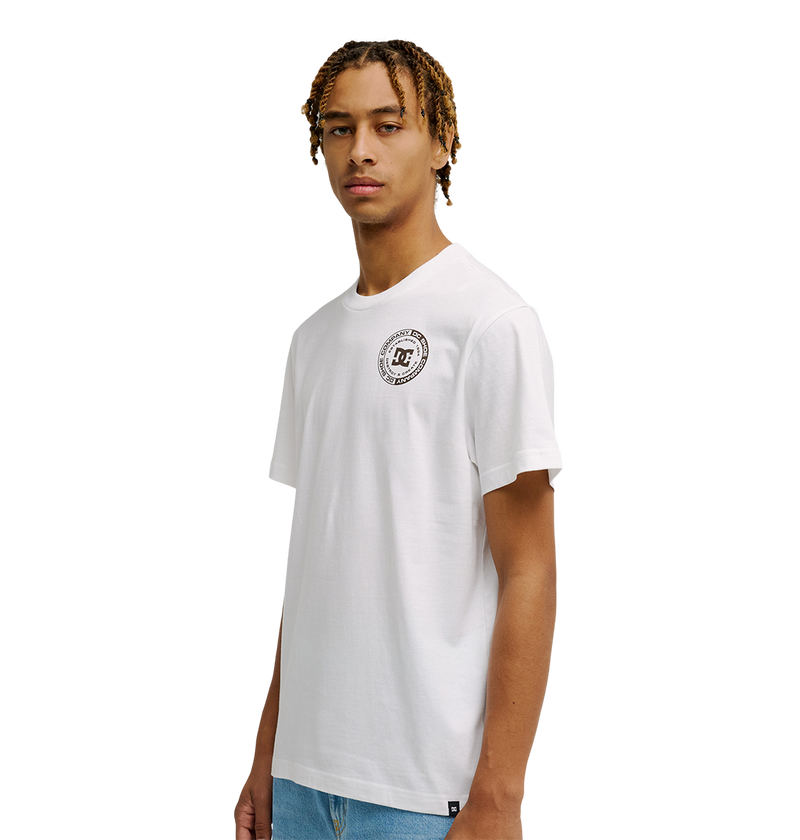 A young man with short, twisted hair models the Men's DC Corpo Tee by DC Shoes—a white relaxed fit t-shirt with a corporate logo—paired with light blue jeans. He stands against a plain white background, facing the camera.