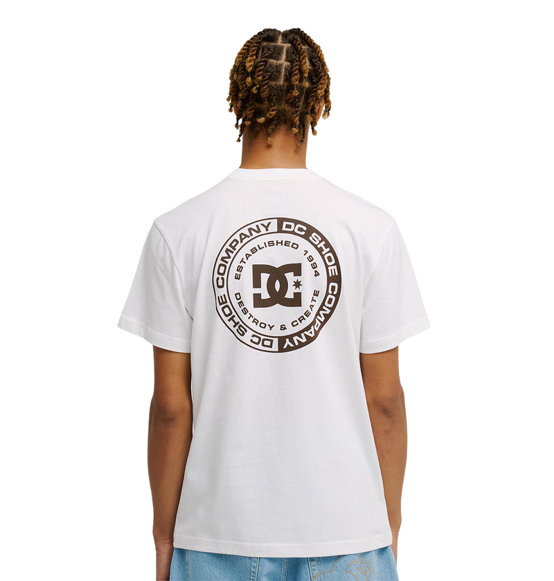 A person faces away wearing the Men’s DC Corpo Tee by DC Shoes, featuring a large circular skate-inspired logo and black text on the back, paired with light blue jeans. The background is plain white.