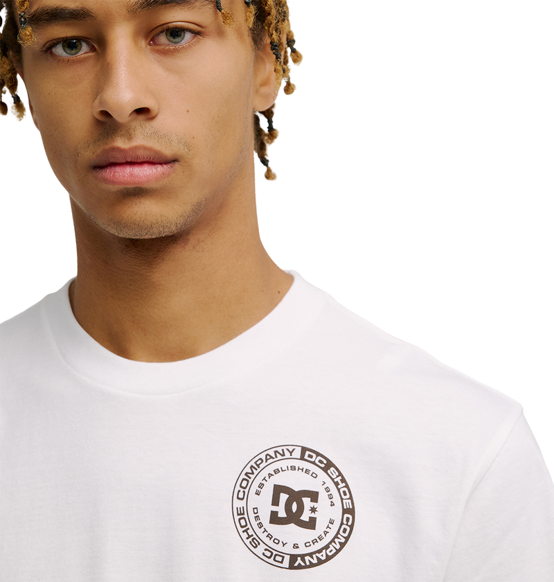 A young man wears the Men's DC Corpo Tee by DC Shoes, a relaxed fit white skate t-shirt with a circular corporate logo and surrounding text on the upper left chest. The background is plain white.