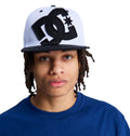 A person with shoulder-length dreadlocks wears a blue shirt and a white and black Reebok DC Double That Cap, gazing at the camera against a plain white background.