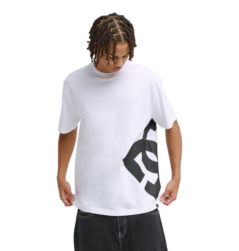 A young man with braided hair wears the Reebok DC Lanai Hss, a loose white T-shirt with a bold black abstract side design, paired with dark jeans. He looks down and holds the hem of his shirt against a plain white background.