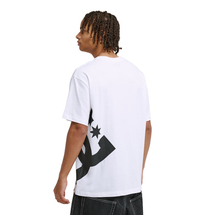 A person with short, twisted hair models the Reebok DC Lanai Hss white t-shirt with a bold black star and curved graphic on the back, standing side and back to the camera against a plain white background.