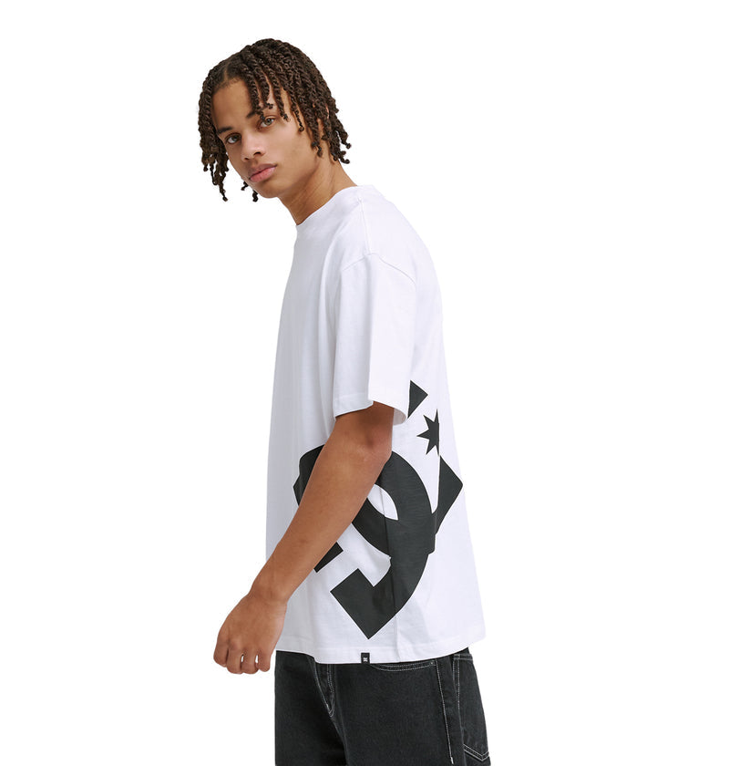 A person with short, twisted hair wears the Reebok DC Lanai Hss, a loose white t-shirt with a bold black graphic on the side, paired with black jeans, standing against a plain white background and looking over their shoulder at the camera.