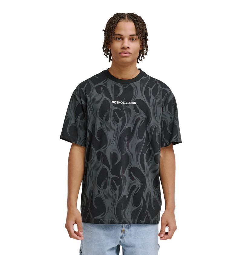 A young person with short, curly hair wears the Reebok DC Liquid Fuego Hss t-shirt—black with a gray flame pattern and "Reebok" printed in white on the chest—paired with light blue jeans, standing against a plain white background.