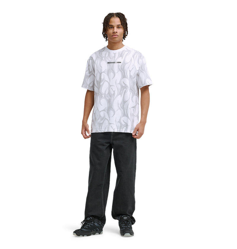 A person with short, braided hair wears the Reebok DC Liquid Fuego Hss white patterned t-shirt with text on the chest, paired with black jeans and black sneakers, standing against a plain white background.
