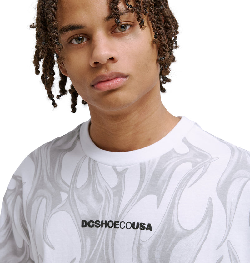 A person with short, curly hair wears the Reebok DC Liquid Fuego Hss white t-shirt with gray flame patterns and DCSHOECOUSA text in black on the chest, looking directly at the camera.