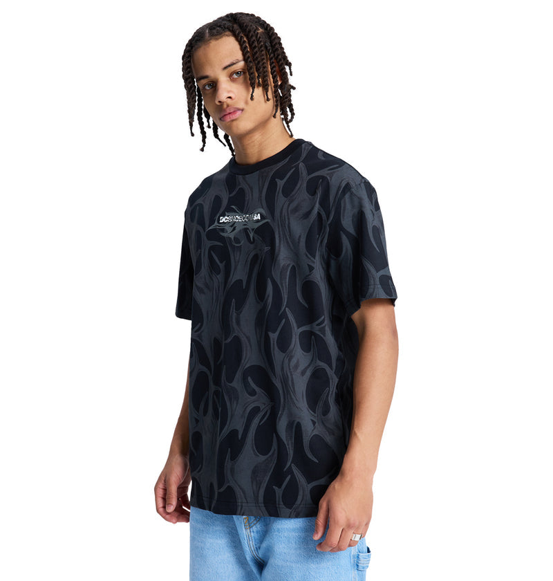 A person with medium-length braided hair models the Reebok DC Liquid Fuego Hss t-shirt, featuring a gray flame pattern on black, paired with light blue jeans against a plain white background.