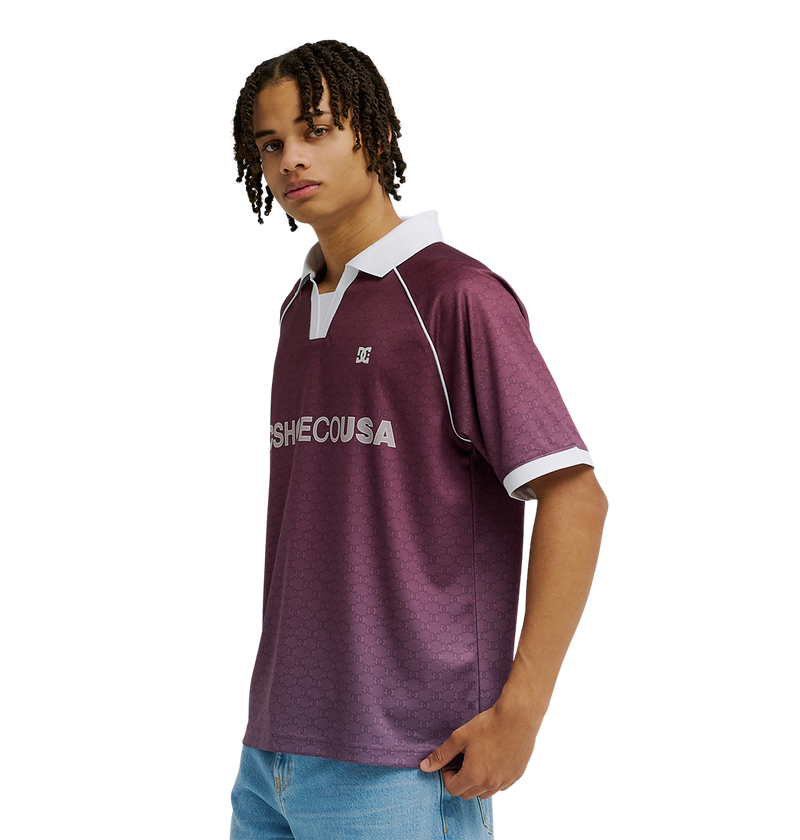 A man with short, curly hair wears the Men's DC Soccer Top by DC Shoes—a maroon polo with white collar and sleeve trim, athletic styling, and breathable fabric—paired with light blue jeans, standing against a plain white background.