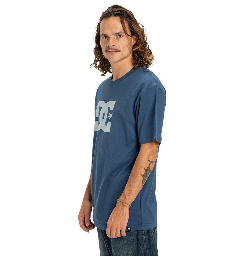 A man with long wavy hair and a mustache wears the Men's DC Star Tee by DC Shoes, showing his right forearm tattoo and gazing at the camera with a neutral expression against a white background.