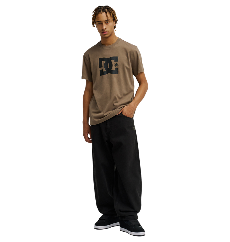 A person with short blonde dreadlocks wears a relaxed fit Men's DC Star Tee by DC Shoes in brown, paired with loose black pants and black sneakers, standing against a plain white background with one hand in their pocket.
