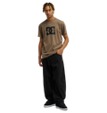 A person with short blonde dreadlocks wears a relaxed fit Men's DC Star Tee by DC Shoes in brown, paired with loose black pants and black sneakers, standing against a plain white background with one hand in their pocket.