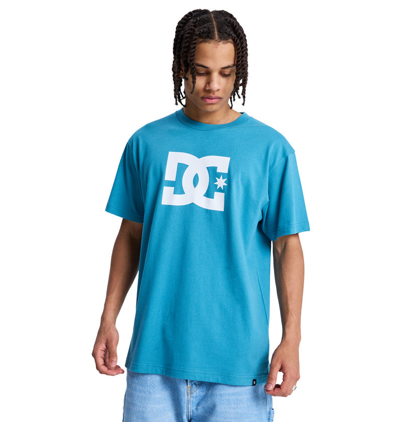 A person with braided hair wears the Reebok DC Star Hss, a loose turquoise T-shirt with a large white logo, paired with light blue denim shorts against a plain white background, looking down.