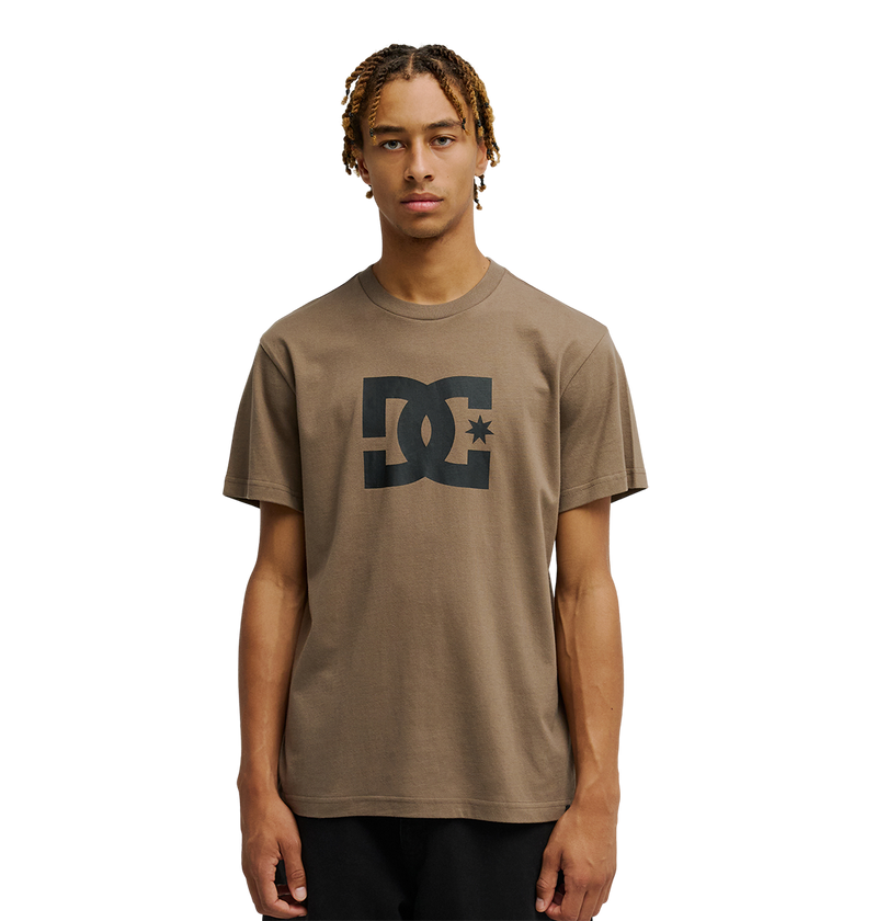 A young man with short, curly blond hair wears the Men's DC Star Tee by DC Shoes, a relaxed fit brown shirt with bold black DC artwork on the chest. He stands facing forward against a plain white background.