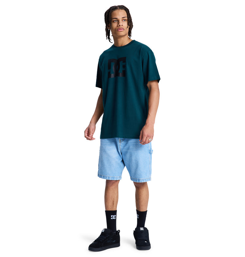 A person with braided hair wears a dark green Reebok DC Star Hss t-shirt, light blue denim shorts, black socks, and black sneakers while standing against a plain white background.