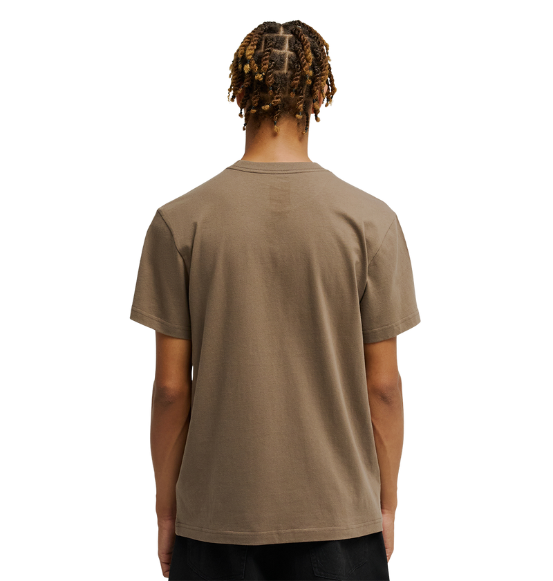 A person with short, twisted hair faces away, wearing the Men's DC Star Tee by DC Shoes in taupe and black pants against a white background.