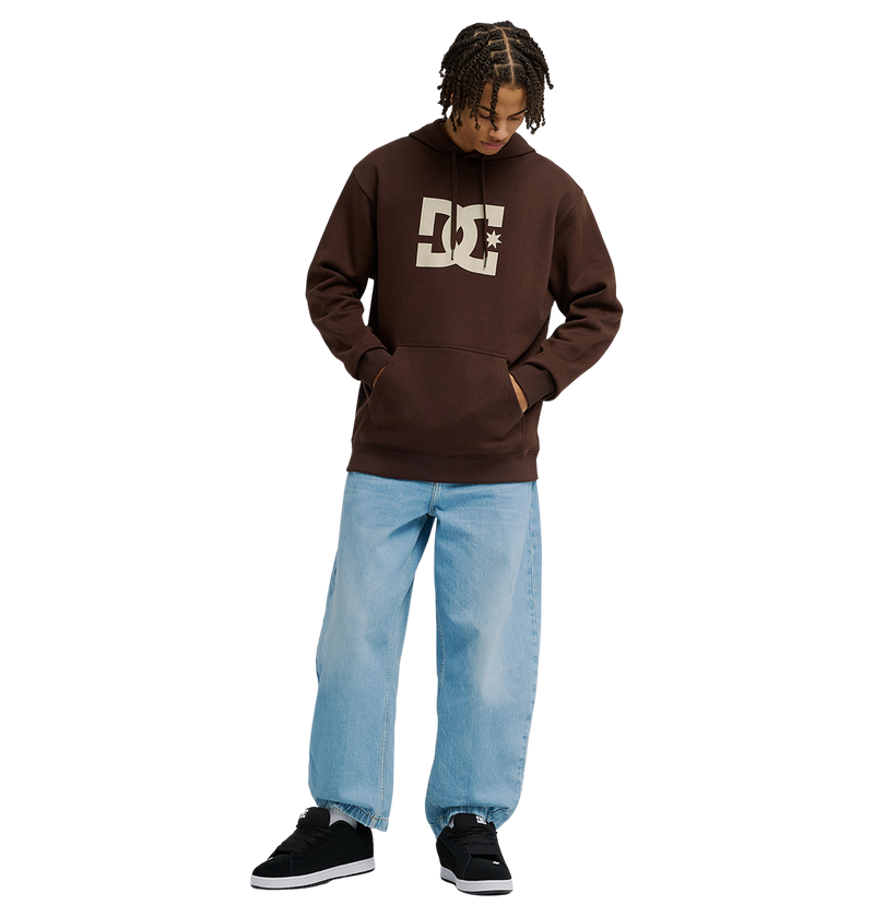 A man wears a brown Men's DC Star Hoodie by DC Shoes, paired with light blue baggy jeans and black sneakers, standing against a plain white background with hands in the hoodie pocket and looking down.
