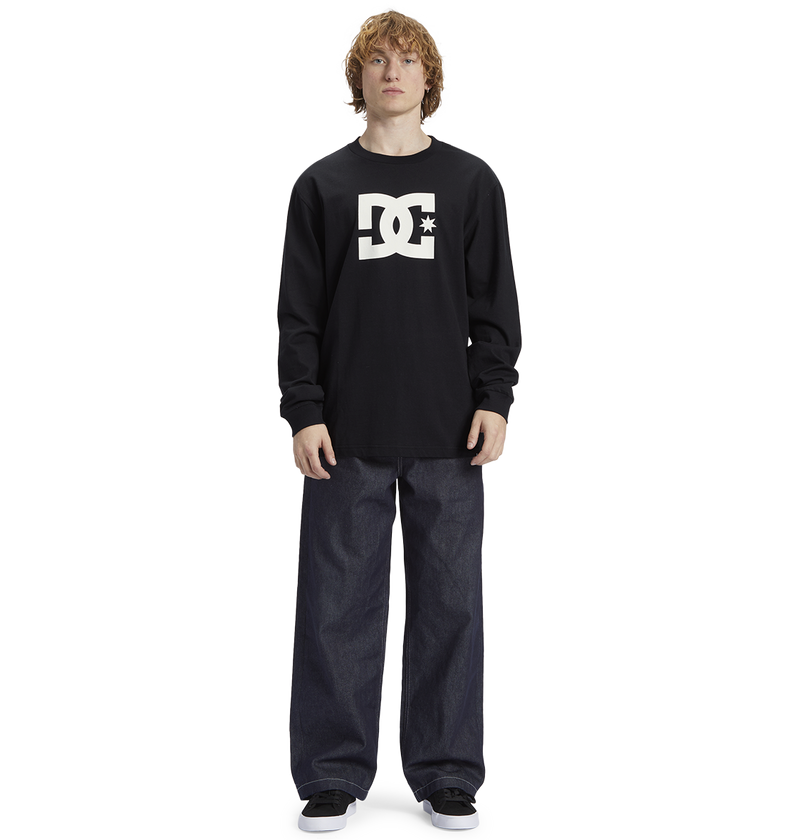 Young person with curly hair wears a black Men's DC Star Tee by DC Shoes, featuring bold graphics, paired with loose dark jeans and black sneakers, standing against a white background—ready to skate.