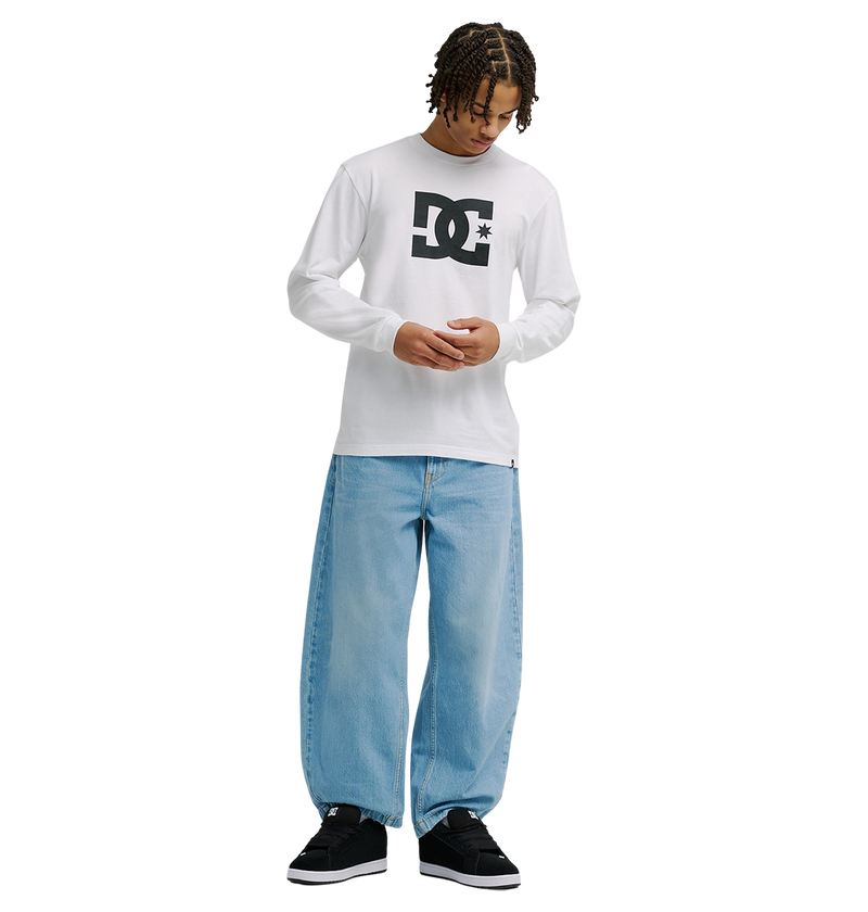 A person with short dreadlocks wears the Men's DC Star Tee by DC Shoes—a white long-sleeve shirt with bold black DC artwork—paired with loose light-blue jeans and black sneakers, standing against a plain white background looking down.
