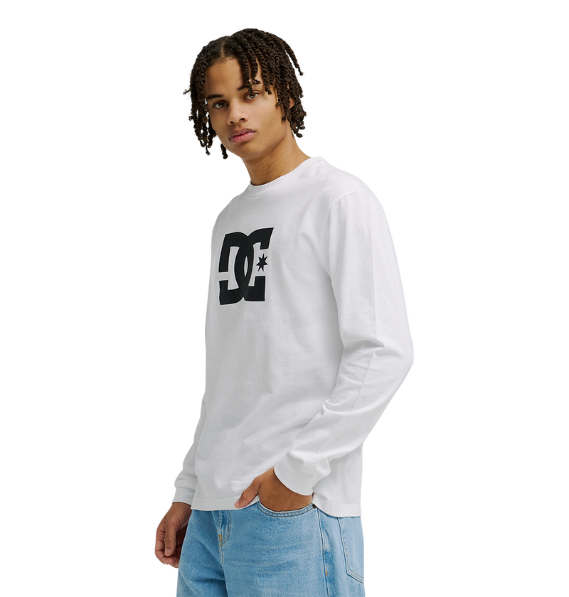 A person with short curly hair wears the Men’s DC Star Tee by DC Shoes, a white long-sleeve graphic shirt with a prominent black logo, paired with light blue jeans, standing against a plain white background.
