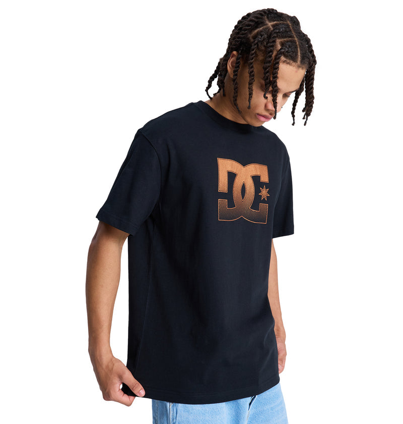 A person with braided hair wears a black Reebok DC Star Oxidized Hss t-shirt featuring a large bronze DC logo on the chest and light blue jeans, looking down against a plain white background.