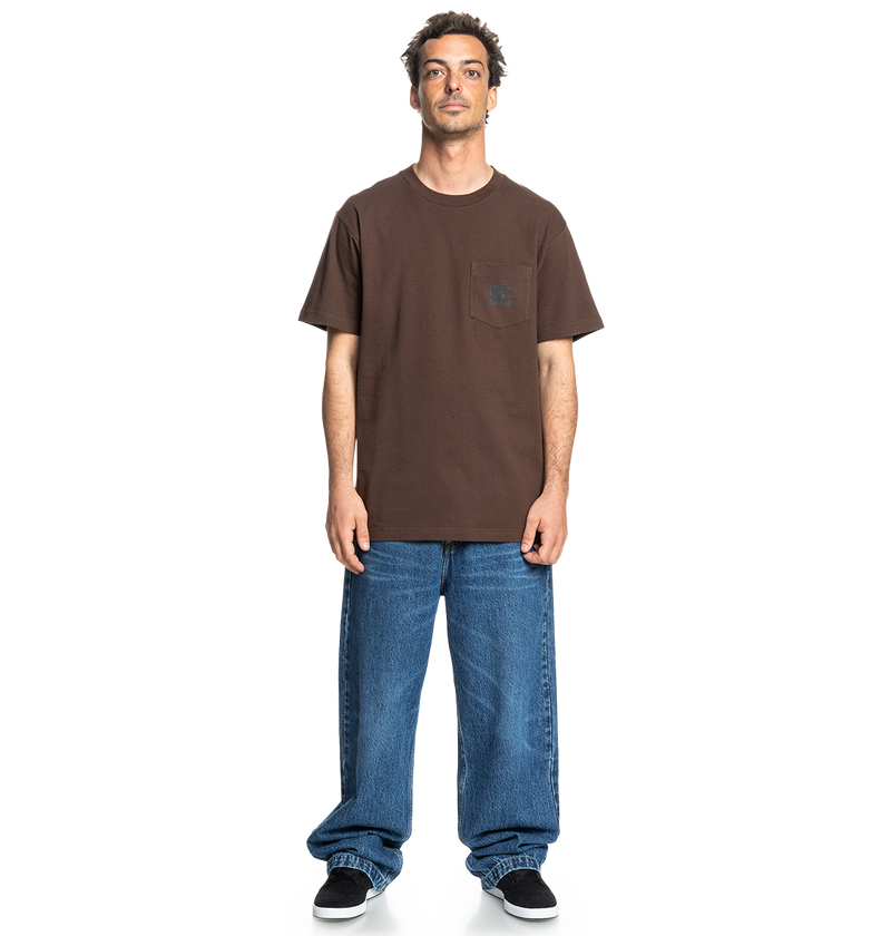 A man faces forward in a relaxed fit brown Men's DC Star Pocket Tee by DC Shoes, featuring a chest pocket, paired with loose blue jeans and black skate shoes against a plain white background.