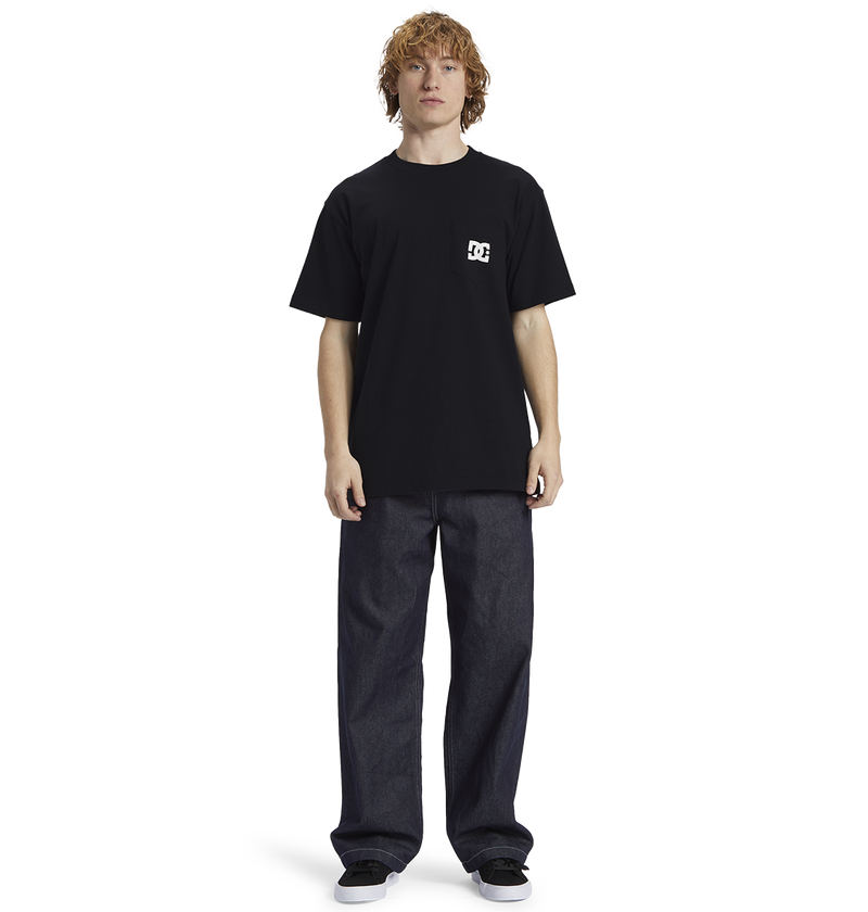 A person with curly hair wears the Men's DC Star Pocket Tee by DC Shoes in black, paired with relaxed fit dark jeans and black shoes, standing against a plain white background.